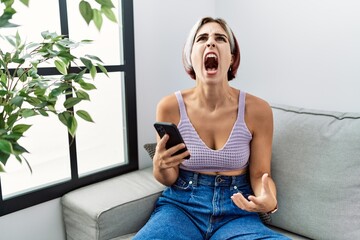 Young beautiful woman using smartphone typing message sitting on the sofa angry and mad screaming...