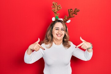 Middle age caucasian woman wearing cute christmas reindeer horns looking confident with smile on face, pointing oneself with fingers proud and happy. © Krakenimages.com