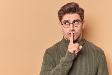 Be quiet. Serious mysterious man presses index finger to lips shows taboo sign tells to keep silence shushing indoor wears casual clothes looks away isolated over beige background blank copy space - obrazy, fototapety, plakaty