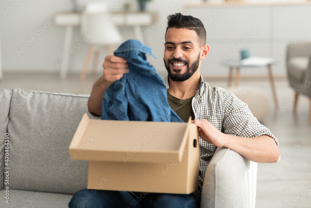 Wall mural Happy millennial Arab guy unboxing carton parcel, satisfied with successful shopping, taking out new clothes at home