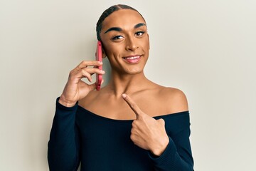 Hispanic transgender man wearing make up and long hair having conversation talking on the smartphone smiling happy pointing with hand and finger