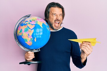 Fototapeta premium Middle age handsome man holding paper plane and world ball angry and mad screaming frustrated and furious, shouting with anger. rage and aggressive concept.