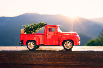 Red retro small car carrying christmas tree in the mountain in the sunbeams of sunset. Winter holiday concept