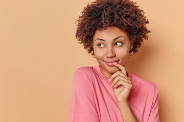 Fototapeta na wymiar Portrait of good looking curly haired young woman looks left with dreamy expression smiles gently wears casual clothes admires something isolated over beige background with blank copy space.