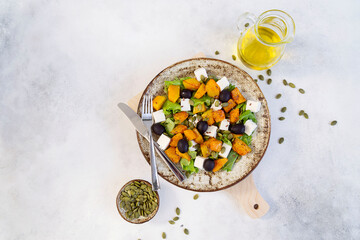 Healthy salad with fried pumpkin, feta cheese, olives, salad greens, pumpkin seeds, olive oil on the light grey background.