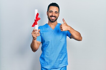 Handsome hispanic man with beard wearing blue male nurse uniform holding diploma smiling happy and...