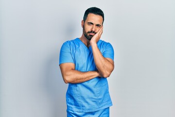 Handsome hispanic man with beard wearing blue male nurse uniform thinking looking tired and bored with depression problems with crossed arms.