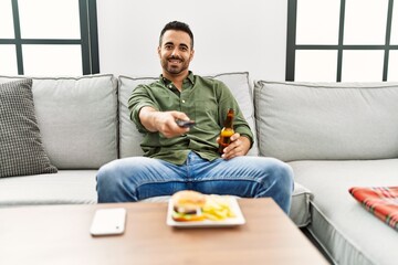 Young hispanic man eating hamburger watching tv at home