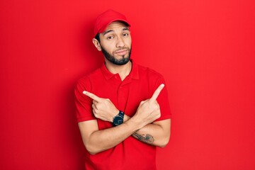 Hispanic man with beard wearing delivery uniform and cap pointing to both sides with fingers, different direction disagree