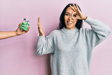 Young hispanic girl rejecting jar of pills with hand smiling happy doing ok sign with hand on eye...