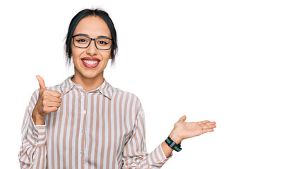 Young hispanic girl wearing casual clothes and glasses showing palm hand and doing ok gesture with thumbs up, smiling happy and cheerful