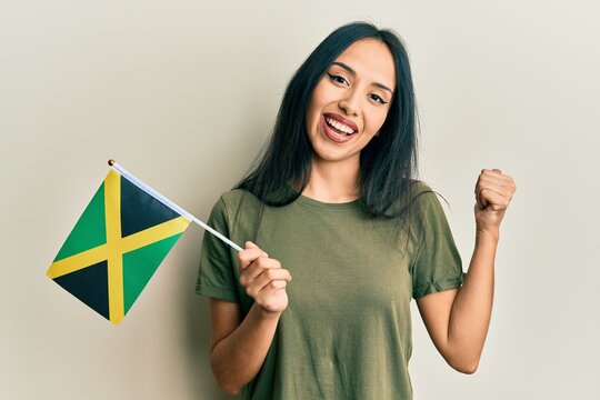 Young hispanic girl holding jamaica flag pointing thumb up to the side smiling happy with open mouth