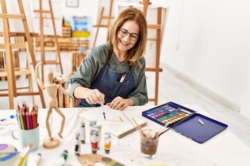 Middle age artist woman smiling happy painting sitting on the table at art studio.