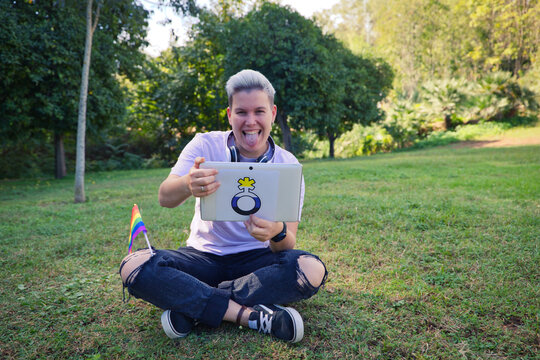 non-binary person sitting in the park with a tablet with the non-binary symbol and the gay pride flag on his knee. Diversity and gay pride. Non-binary visibility concept.