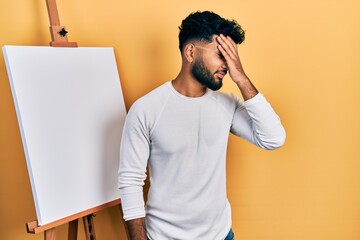 Arab man with beard standing by painter easel stand surprised with hand on head for mistake,...