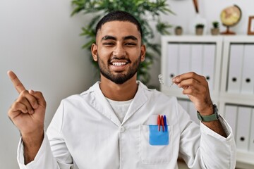 Young indian man working at dentist clinic holding invisible aligner smiling happy pointing with hand and finger to the side