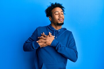 Young african american man with beard wearing casual sweatshirt smiling with hands on chest, eyes...