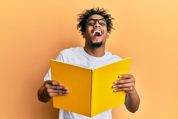 Young african american man with beard reading a book wearing glasses smiling and laughing hard out...