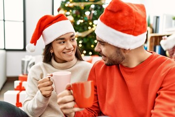 Young hispanic couple smiling happy and drinking cup of coffee sitting on the floor by christmas tree at home.