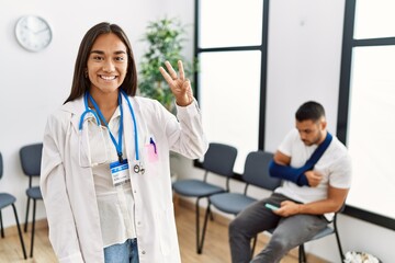 Young asian doctor woman at waiting room with a man with a broken arm showing and pointing up with fingers number three while smiling confident and happy.