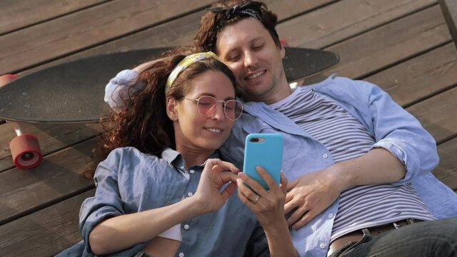 Top view of man and woman with skateboard watching video or photos on smartphone. Smiling boyfriend and girlfriend lying on wooden surface and enjoying time together. Relationship, entertainment idea