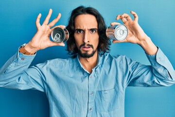 Young hispanic man holding canned food over eyes clueless and confused expression. doubt concept.