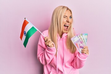 Young blonde woman holding india flag and rupee banknotes angry and mad screaming frustrated and furious, shouting with anger. rage and aggressive concept.