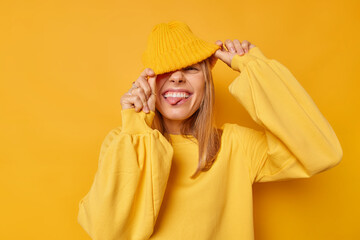 Joyful happy woman hides eyes with hat sticks out tongue has playful expression teases someone wears casual jumper isolated over yellow background squints happily shows true emotions. People and fun