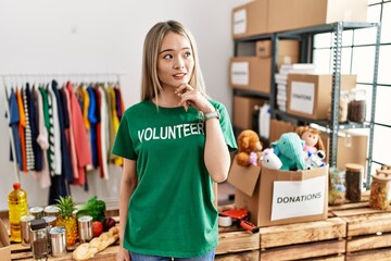 Asian young woman wearing volunteer t shirt at donations stand with hand on chin thinking about question, pensive expression. smiling and thoughtful face. doubt concept.