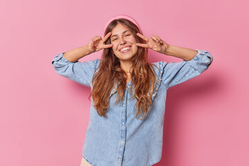 Young happy woman sends positive vibes makes peace gesture over eyes smiles toothily dressed in denim shirt isolated over pink background shows victory sign has cheerful mood. Body language.