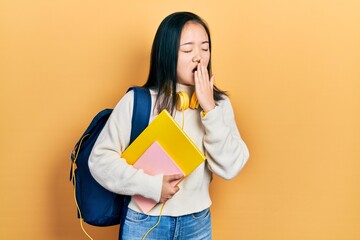 Young chinese girl holding student backpack and books bored yawning tired covering mouth with hand....