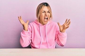 Young caucasian woman wearing casual clothes sitting on the table crazy and mad shouting and...