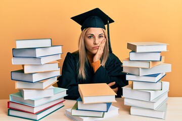 Young caucasian woman wearing graduation ceremony robe sitting on the table thinking looking tired and bored with depression problems with crossed arms.