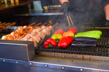 A male chef fries meat and various vegetables on the grill. Close-up on food and hands of a cook. Cook in black apron fries on fire. Barbecue in open air.