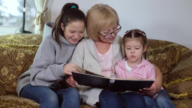 Elderly mature woman is a grandmother, looking at a family photo album with her granddaughters sitting on the sofa, remembering stories from the past. The concept of history and biography.