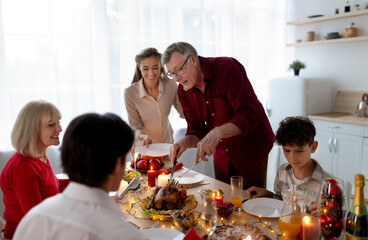 Positive senior man cutting tasty roasted turkey, celebrating Thanksgiving or Christmas with his big extended family