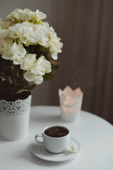 A cup of coffee, a pot with a white flower and a burning candlestick stand on a white table. The coffee is hot, there is steam