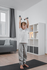 Fototapeta na wymiar Sports hall at home. Boy child exercising with dumbbells in the living room. The child is dressed in a white t-shirt and gray pants.