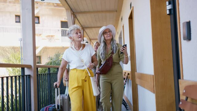 Senior women friends with suitcases standing in front of hotel room outdoors on terrace.
