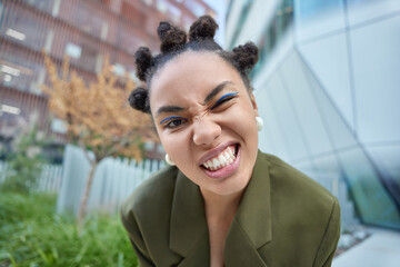 Close up shot of cheeky dark skinned woman winks eye clenches teeth wears blue eyeliner dressed in...