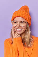 Pleased young pretty woman keeps hands near face smiles gently with teeth looks away wears orange long sleeved jumper and hat isolated over purple background has pleasant thoughts. Happy emotions