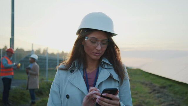 Caucasian young pretty woman engineer wearing hardhat using mobile phone on solar field construction. Power plant workers. Professional people. Technology.