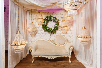 Elegant photozone decorated with sofa, lamps and greenery wreath on background of white brick wall. Photo booth on luxury event. Decor on wedding or birthday party.