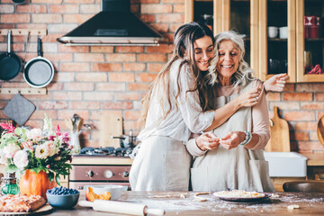 Young woman and mother in white aprons stand at large brown wooden kitchen table and prepare tasty dinner. Senior mother and happy adult daughter baking pumpkin pie together at home. - obrazy, fototapety, plakaty