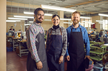 Portrait of three happy workers standing in a workshop at a shoe or garment factory. Two men and a woman in aprons smiling looking at the camera. Footwear and apparel manufacturing industry concept.