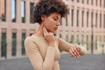 Athletic young woman controls pulse after training keeps hand on neck checks heart rate focused at smartwatch dressed in sportswear poses against urban background. Healthy lifestyle concept.