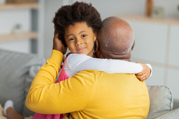 Smiling cute african american little child hugs elderly man on sofa in living room interior