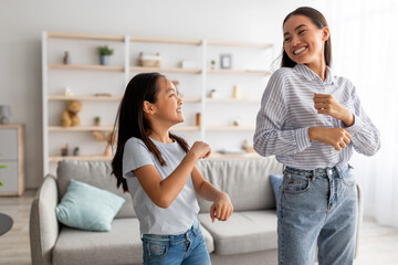 Excited asian mom and cute daughter dancing to music and having fun at home, smiling to each other