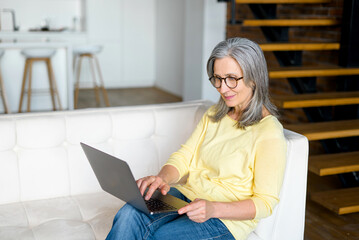 Focused mature woman wearing stylish glasses using a laptop working from home in relaxed atmosphere, charming elderly female concentrated types on the keyboard sitting on the couch