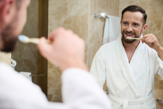 Handsome young bearded attractive European man wearing white terry bathrobe smiles looking at his reflection in the bathroom mirror while brushing teeth